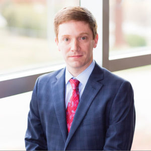 Josh Neighbors wearing a dark blue blazer and red tie.