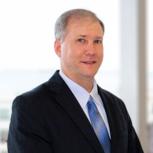 Duane standing front of a window wearing a black suit and blue tie.