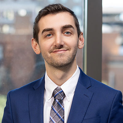 Travis standing in front of a window wearing a blue jacket, white shirt and a blue and gray tie.